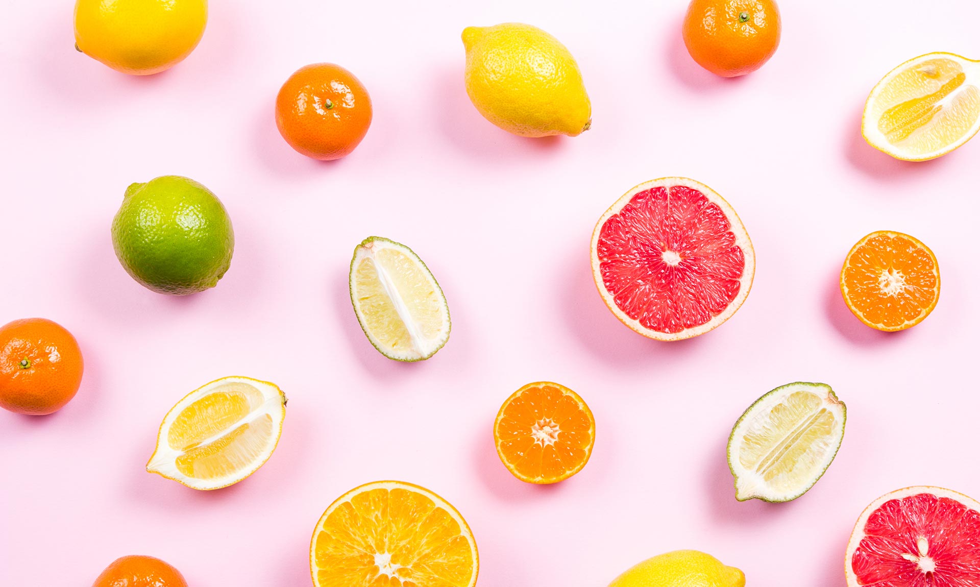 Whole and sliced citrus fruit is scattered against a pink paper backdrop.